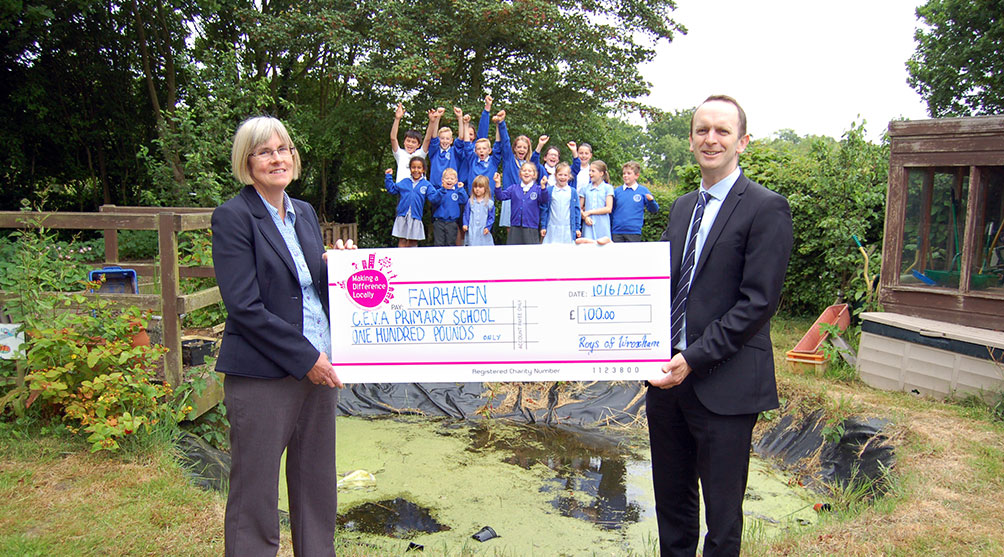 Paul Roy, Buying & Marketing Director presenting a cheque to Head teacher Janice Dix in front of the pond and students from the school